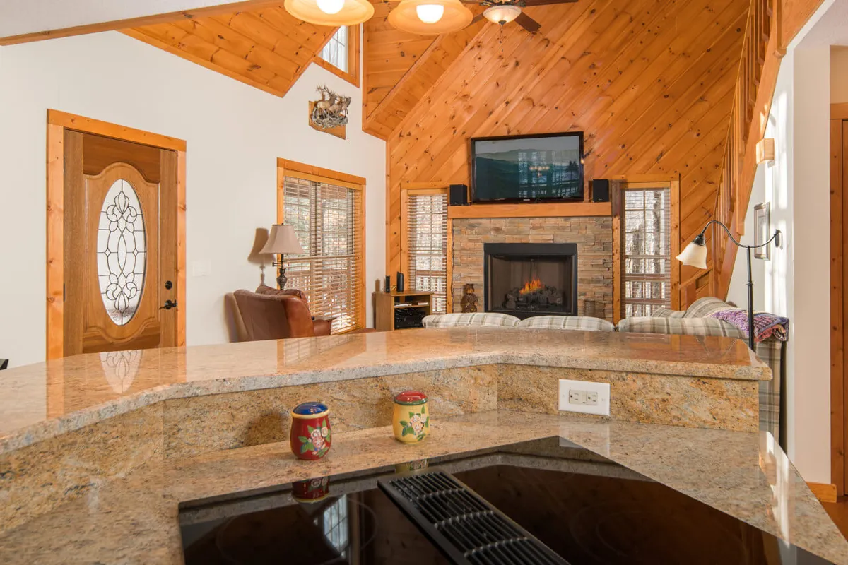 A view from the kitchen area that includes a spacious countertop, modern appliances, a glimpse of the fireplace, large windows with mountain scenery, and exposed wooden beams. The open-concept design seamlessly connects the kitchen, dining, and living areas, inviting group interaction.