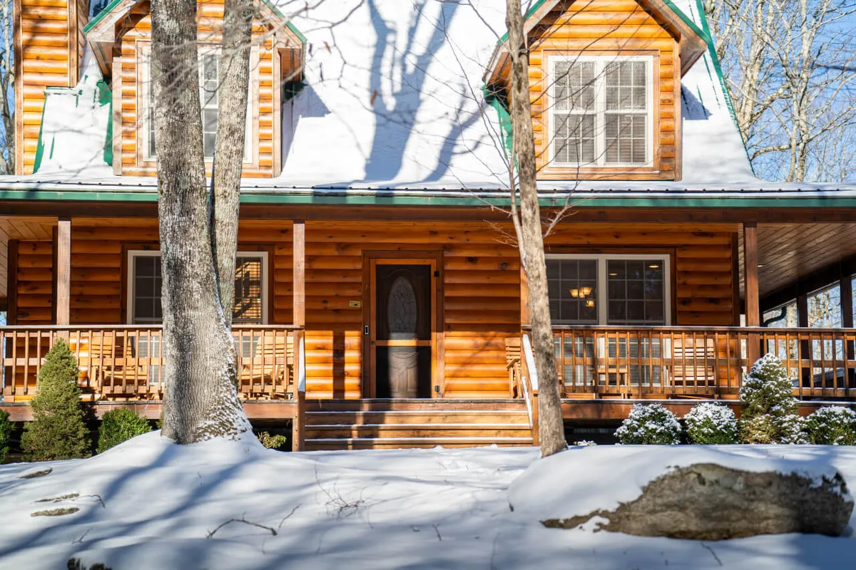 The image captures the wooden log cabin structure with a snow-covered roof and a spacious front porch. There is a wide driveway area visible, suitable for multiple vehicles. Surrounding trees and the yard create a mountain retreat feel that's perfect for families, friends, and pets to arrive and unload gear with ease.