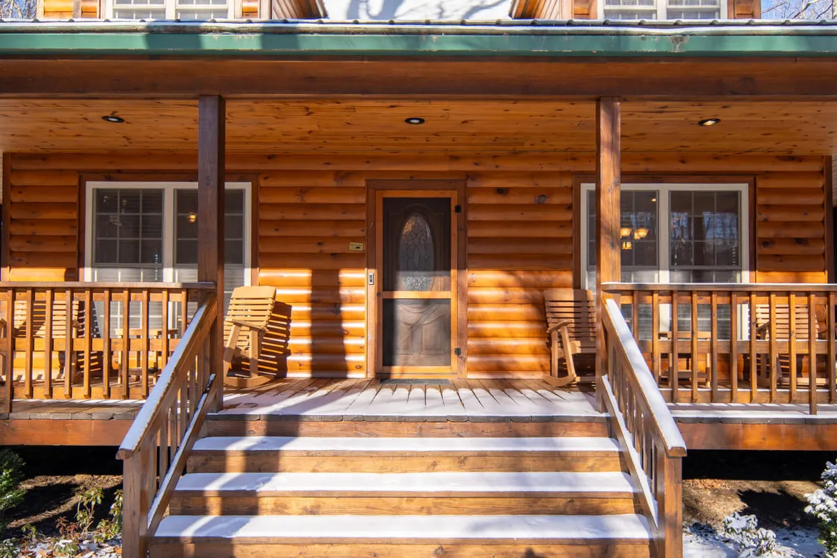 This image shows the wooden porch and front door of Urus Lodge, with stacked chairs and a welcoming entryway. The setting is cozy, with sunrise or evening light highlighting the inviting entrance. The porch provides direct access from the parking area and is free of stairs, making it easily accessible for everyone, including those returning from adventures with pets or gear.