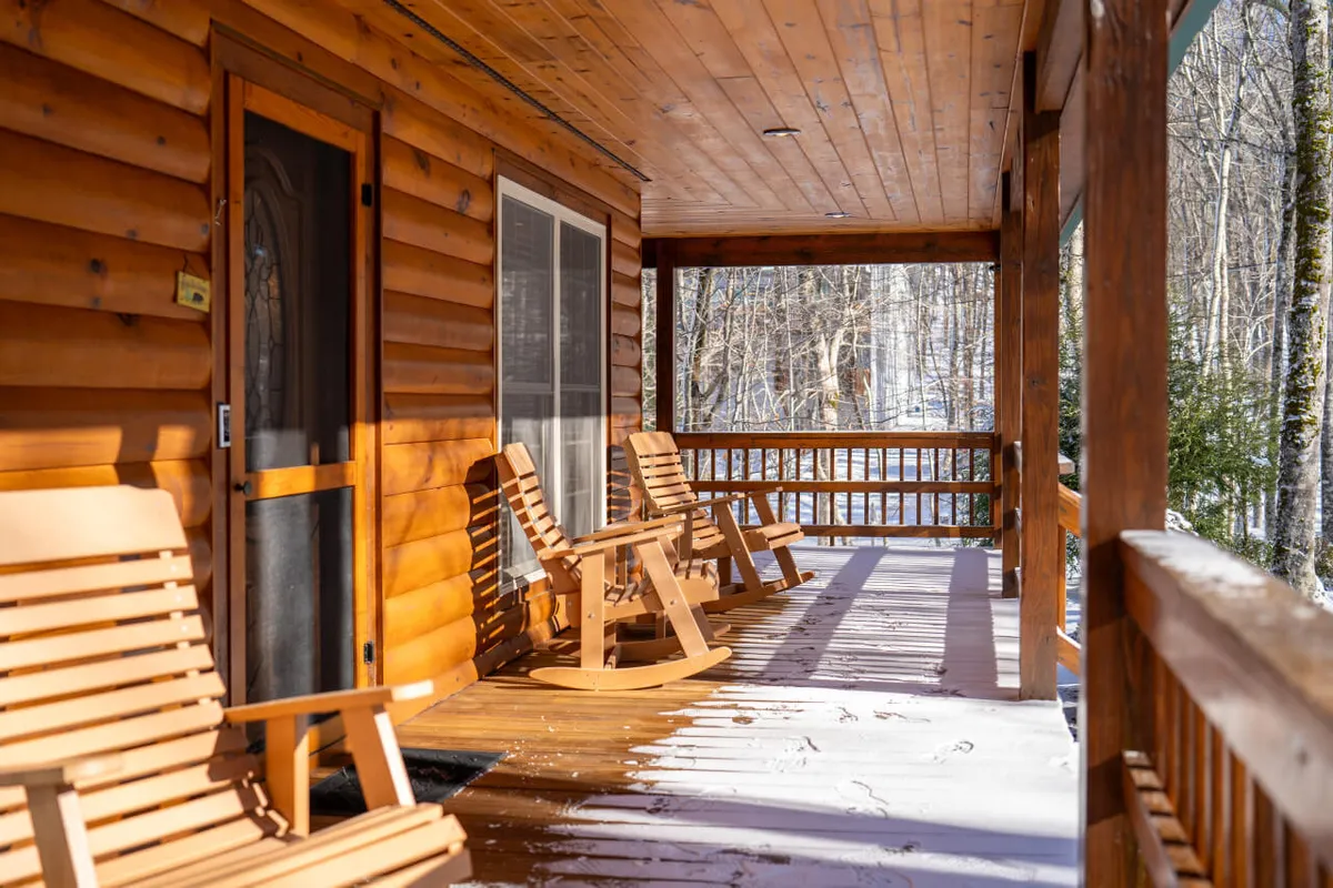 Wooden rocking chairs line a snow-covered porch, inviting guests to relax and take in the snowy scenery. The logs of the cabin and the gentle blanket of snow create an atmosphere of warmth and comfort, even in the chill of winter.