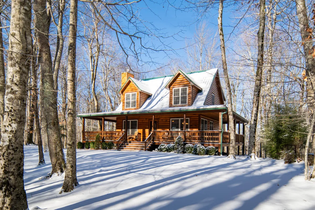 The log cabin stands peacefully amid snow-dusted trees, with the ground transformed into a playground for winter fun. The image features ample space for building snowmen, kids’ activities, and quiet enjoyment of the wintry landscape, making it inviting for families and couples alike.