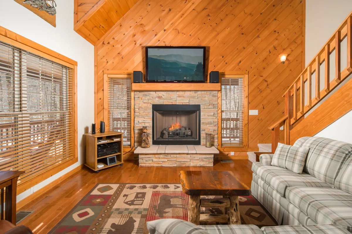 A living room setting with a fireplace, plush sofa, television, and wooden accents. The space is arranged to encourage relaxation and close conversation, with windows that suggest beautiful outdoor vistas.