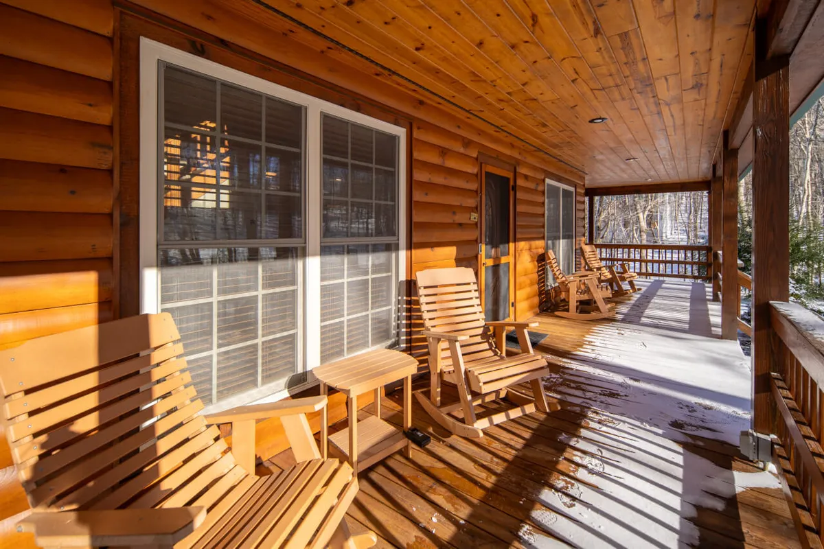 A cozy covered wooden porch wraps around the cabin, featuring classic wooden rocking chairs positioned to take in expansive mountain views. Large windows connect the indoor living spaces with the outdoors, while the inviting mood suggests a perfect spot for relaxing with a book, enjoying morning coffee, or gathering with friends as the sun sets.