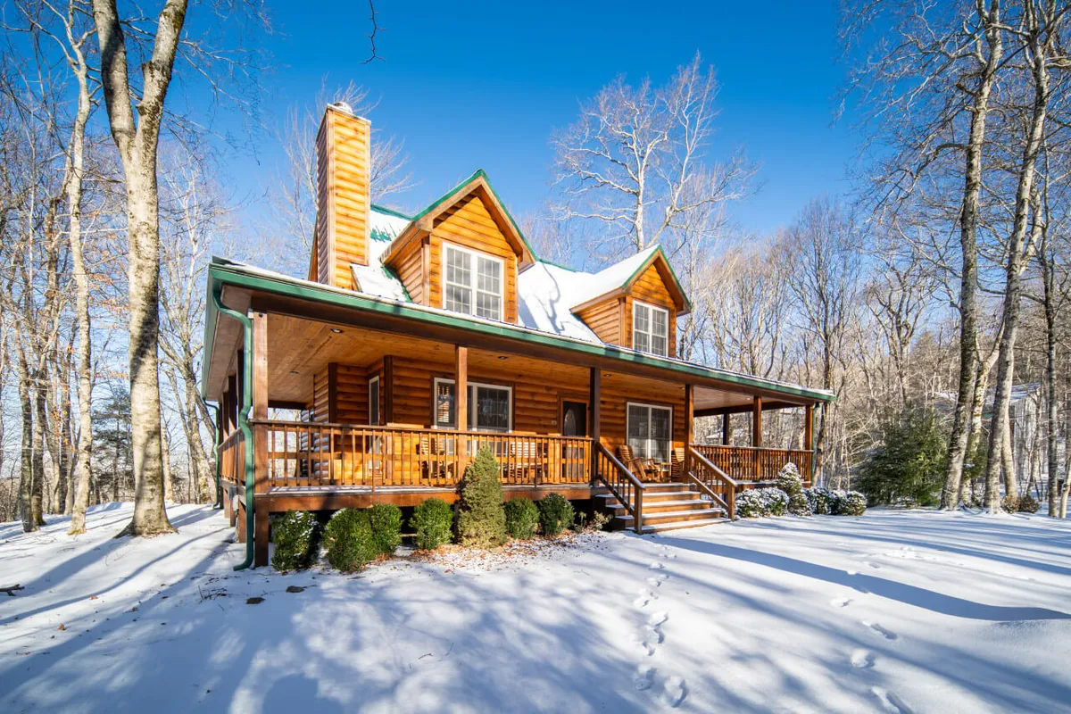 A beautiful log cabin sits nestled among tall trees, its wraparound porch and the surrounding ground blanketed in snow. The tranquil setting evokes a true winter wonderland, with the cabin serving as a cozy base for winter activities. The image hints at the close proximity to both nature and outdoor adventures, ideal for families and groups seeking Beech Mountain's snowy delights.
