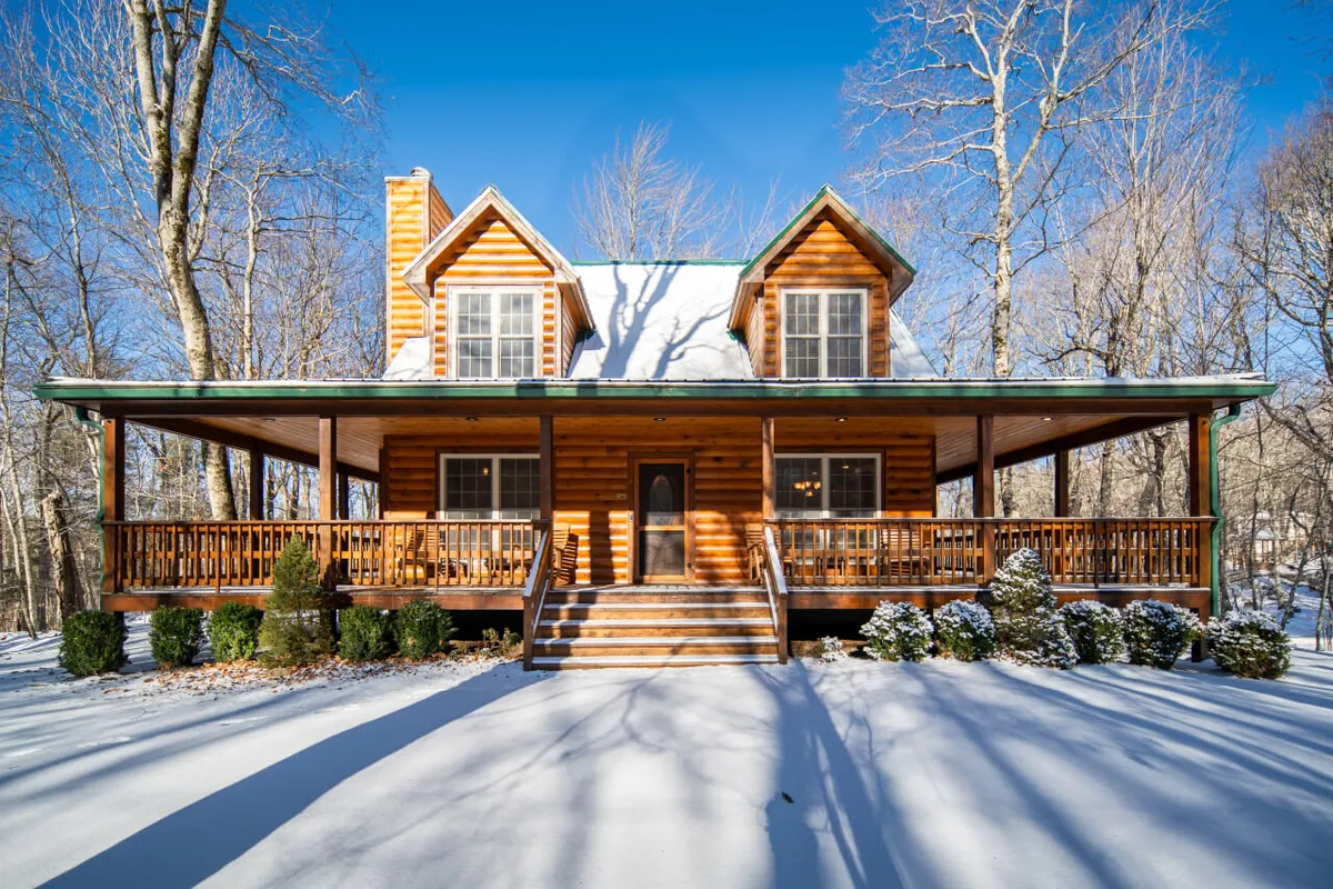The hero image showcases the exterior of a charming log cabin surrounded by snow-dusted trees, with a spacious porch perfect for relaxing and taking in the serene mountain landscape. The inviting glow from the cabin's windows contrasts beautifully with the crisp, snowy ground, evoking a sense of warmth and coziness amidst the winter scenery.