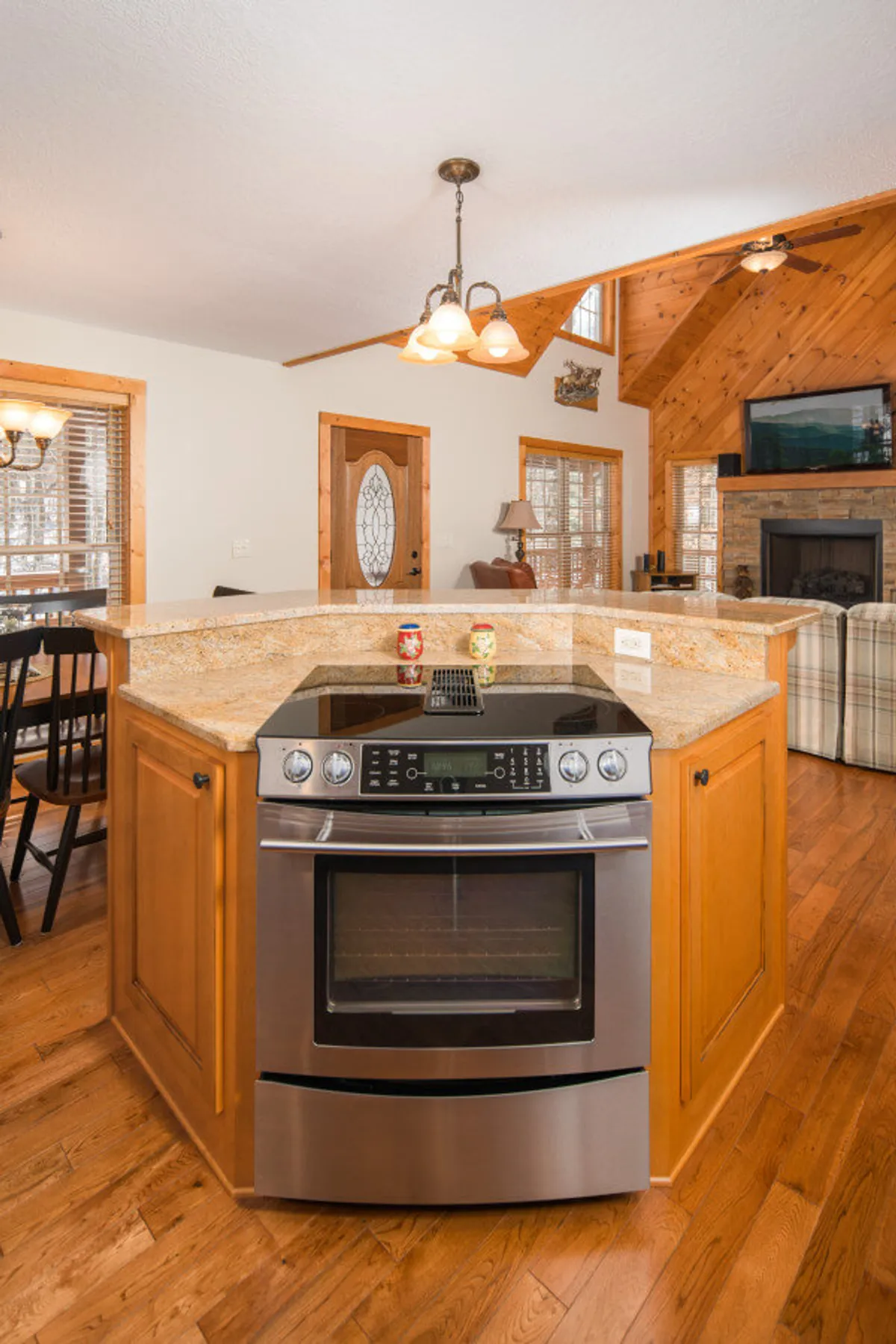 A spacious kitchen and dining area with a stainless steel oven, granite countertops, wooden cabinetry, and a dining table with chairs, all centered around a fireplace. The setup feels welcoming for group meals or celebrations and includes elements that evoke both comfort and togetherness.