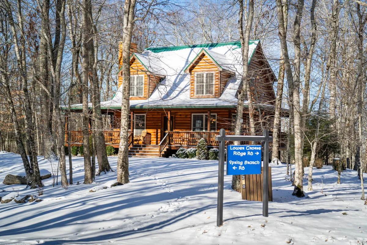 A welcoming exterior shot of a log cabin, nestled among snow-dusted trees. The photo highlights the cabin’s spacious porch with a visible welcome sign, and a wide-open area in the foreground, perfect for outdoor activities, play, or simply soaking up the peaceful mountain surroundings.
