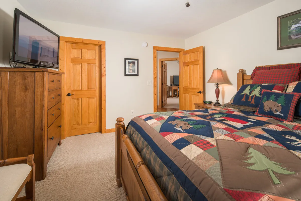 A warm, inviting bedroom featuring a bed with a patchwork quilt, wooden furniture, and a television. The room feels cozy and private, with doorways suggesting en-suite access and natural light filtering in.