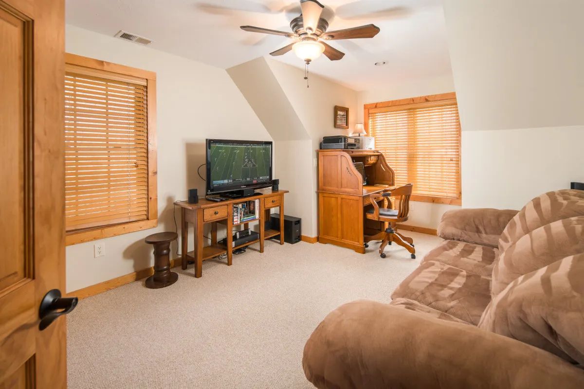 A versatile room featuring a sofa, television, desk with chair, and a window with blinds. The setup is ideal for both relaxation and productivity, offering a comfortable place to work or unwind.