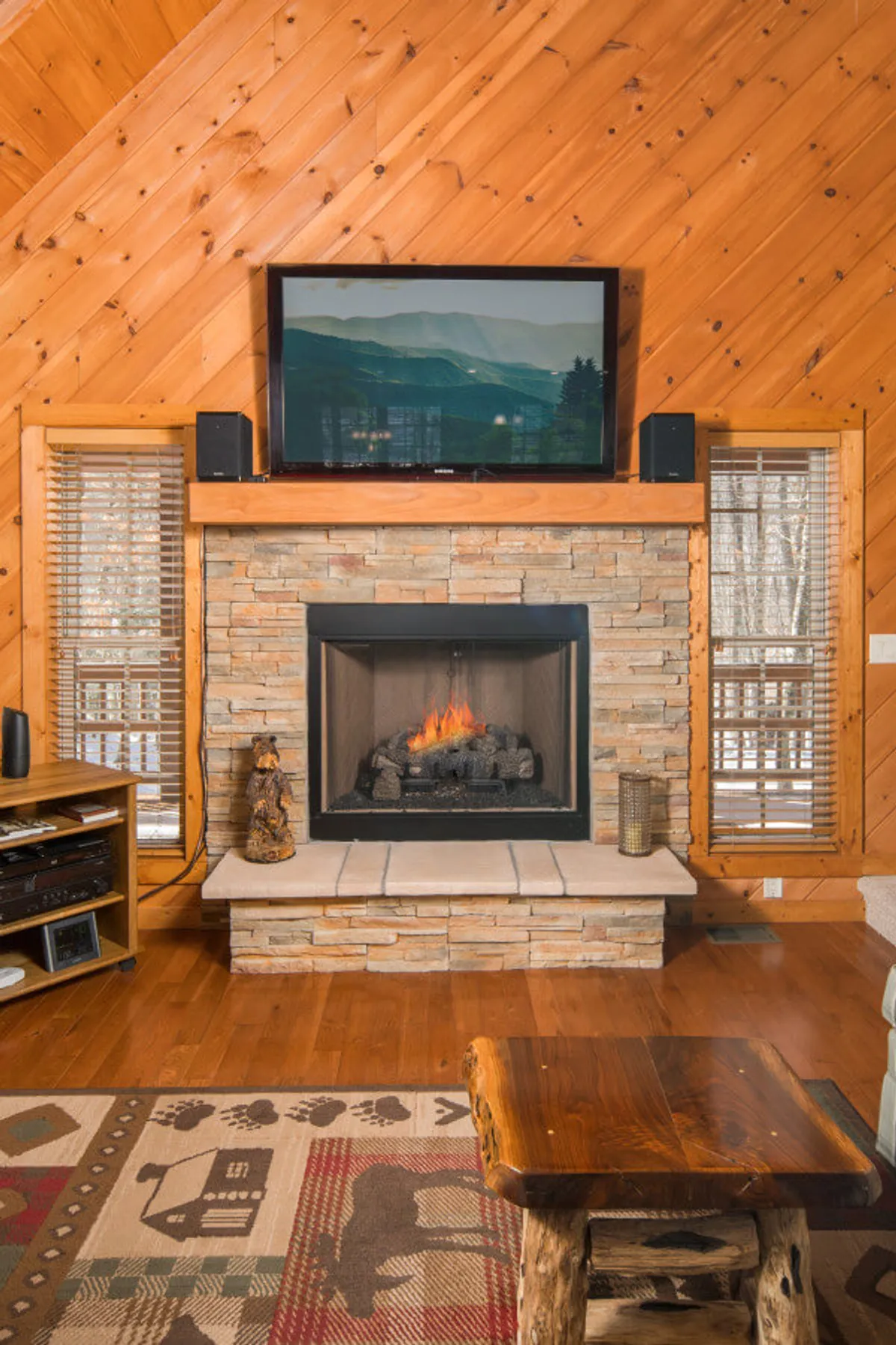 A warmly lit living room featuring a central gas fireplace, a television mounted above, wooden walls, a comfortable sofa, a coffee table, and a rug. Large windows allow natural light to stream in, hinting at scenic mountain views outside. The arrangement creates a cozy and communal gathering space.