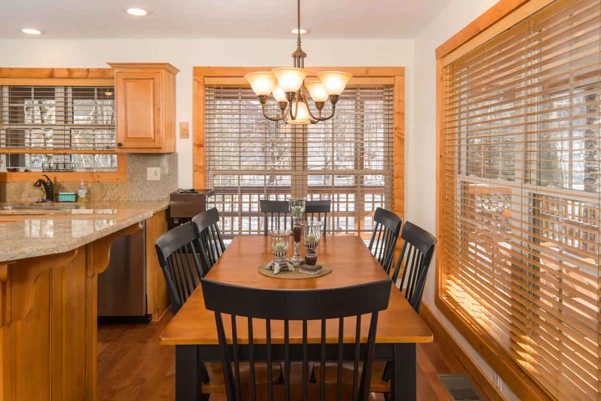 A warmly decorated dining area featuring a large wooden dining table, a sophisticated chandelier above, stylish kitchen cabinetry, and window blinds letting in soft natural light. The ambiance is inviting and ideal for shared meals or celebrations.