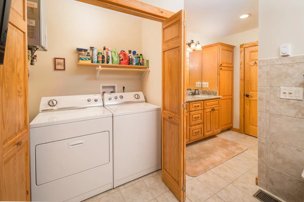 A practical and tidy interior view showing a laundry area with a washing machine, shelf for cleaning products, and wooden cabinetry. This space is useful for organizing and cleaning gear after outdoor adventures, or for pet owners to manage any messes their dogs might bring in from outside.