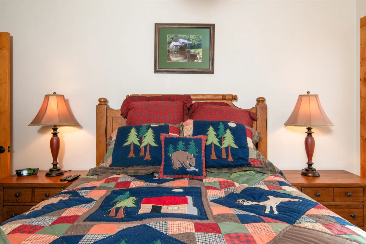 A cozy bedroom with a bed adorned with a patchwork quilt, matching lamps on nightstands, and a framed picture above the bed. The space is inviting, ideal for relaxation and peaceful sleep.